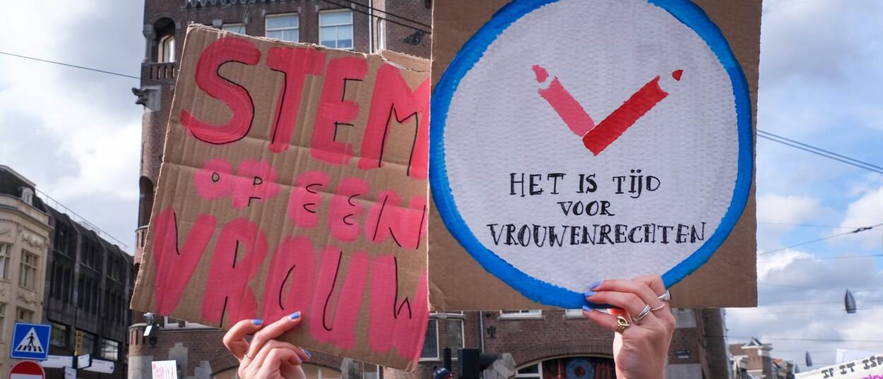 Een vrouw houdt twee zelfgemaakte borden omhoog met daarop de teksten 'stem op een vrouw' en 'het is tijd voor vrouwenrechten'.