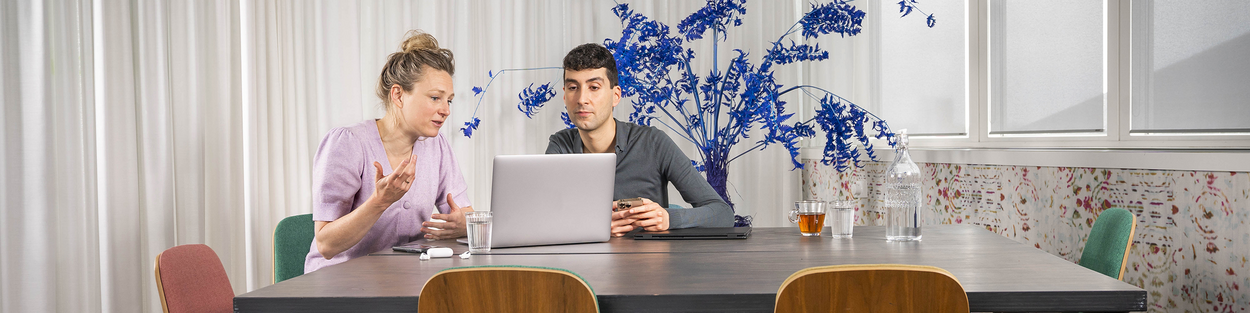 Twee collega's overleggen aan een tafel met daarop twee laptops, een telefoon, een setje oortjes, een kop thee, fles water en een glas