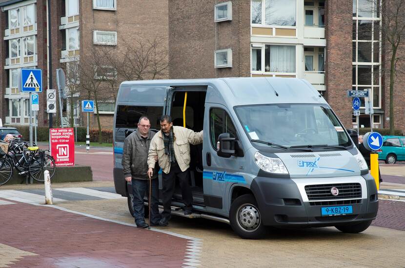 Een man met een wandelstok stapt uit een regiotaxi, de chauffeur helpt de man.