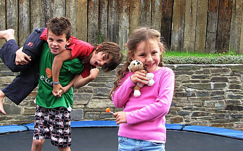 Drie kinderen spelen op een trampoline, de twee jongens zijn aan het stoeien en het meisje speelt met een knuffeltje.